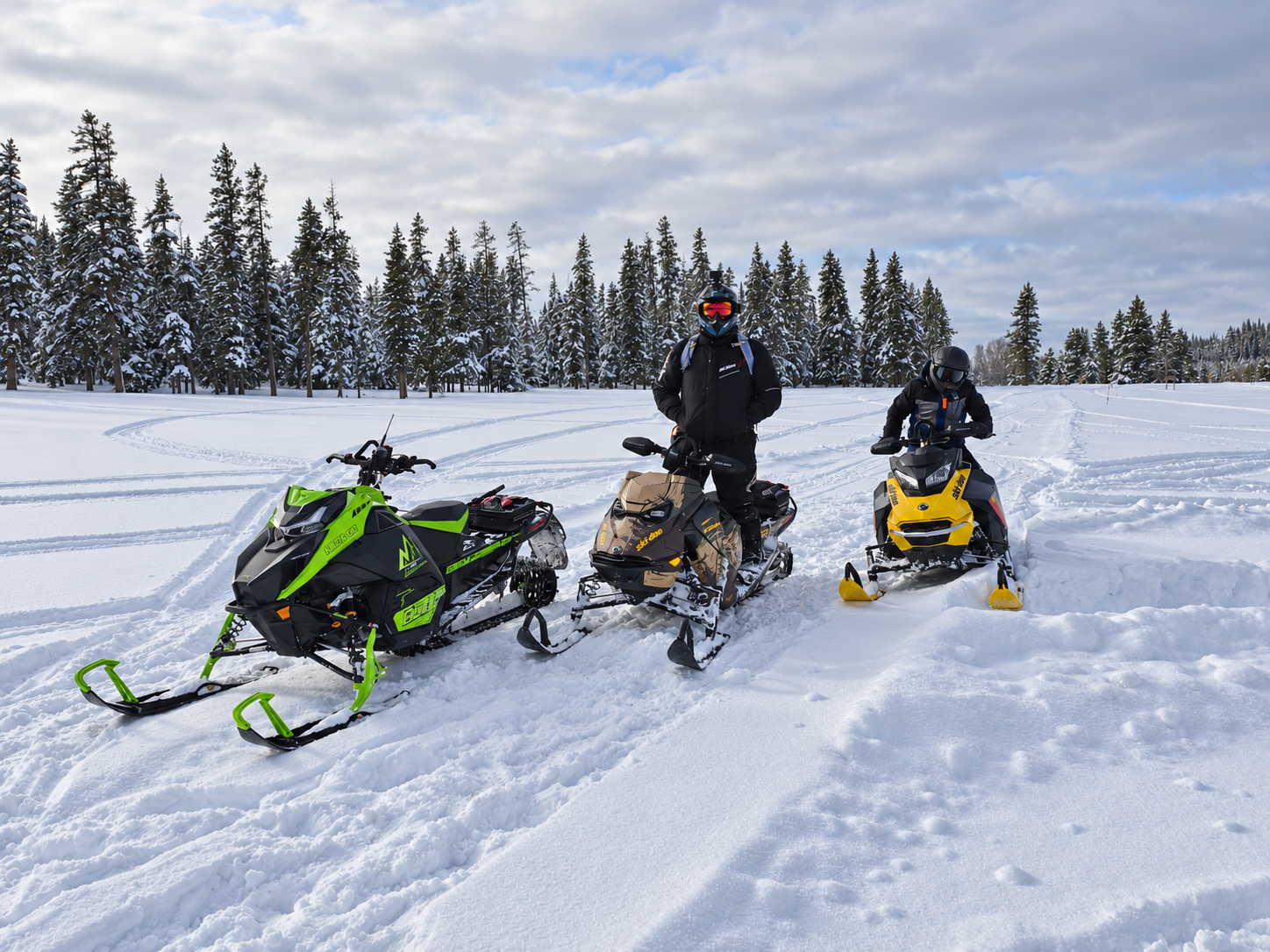 Three snowmobiles and two helmeted riders in a snowy clearing during the 2025–2026 development season